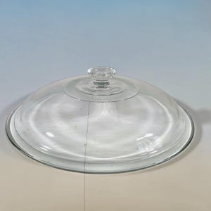 May include: A clear glass dome lid featuring a smooth, rounded surface and a central knob handle. The base of the knob is surrounded by a textured, circular pattern of small, raised dots, providing a decorative detail to the transparent cover.