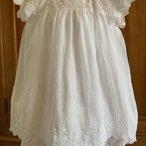 Op de afbeelding: Een witte jurk in vintage-stijl met ingewikkelde kanten details. De jurk heeft een ronde halslijn, korte mouwen en een zwierige rok. Het lijfje en de mouwen zijn versierd met delicate kant, en de zoom is geschulpt.