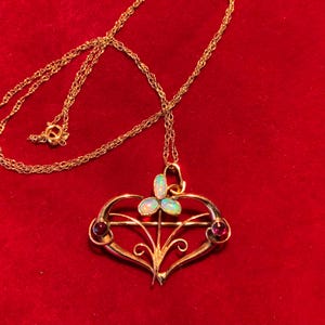 May include: A gold pendant necklace with a heart-shaped design. The pendant has three opalescent stones and two red gemstones. The necklace is displayed on a red velvet surface.