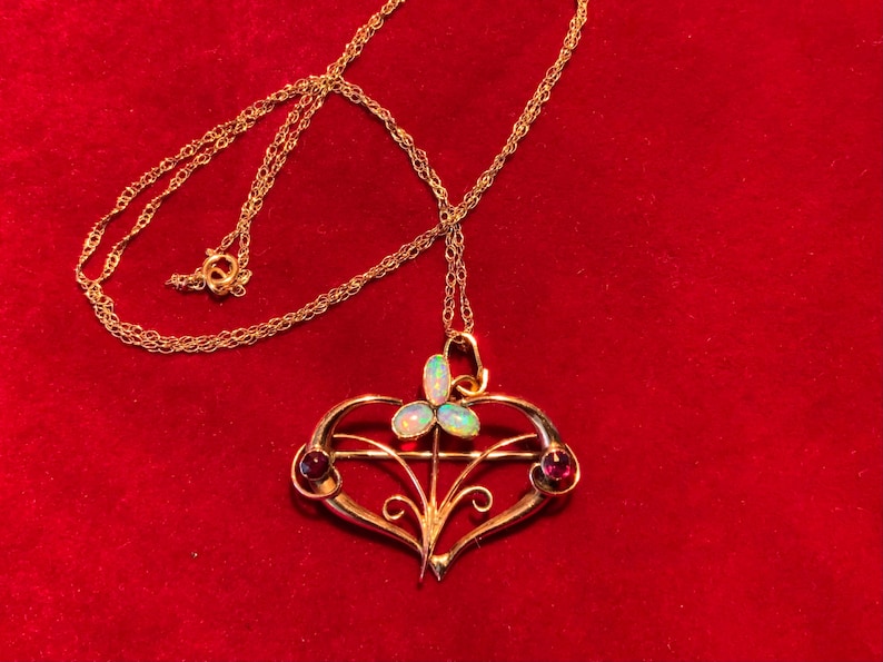 May include: A gold pendant necklace with a heart-shaped design. The pendant has three opalescent stones and two red gemstones. The necklace is displayed on a red velvet surface.