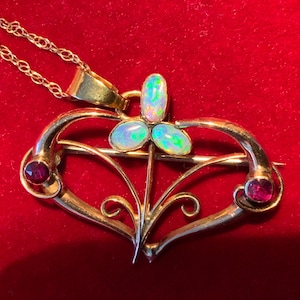 May include: A gold pendant necklace with a heart-shaped brooch design. The brooch features three opal stones and two red gemstones. The necklace is set against a red background.