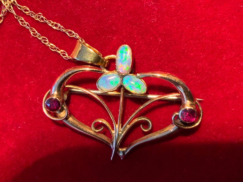 May include: A gold pendant necklace with a heart-shaped brooch design. The brooch features three opal stones and two red gemstones. The necklace is set against a red background.