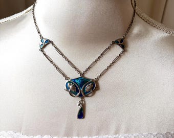 Puede incluir: Un collar de tono plateado con un colgante central que presenta incrustaciones de esmalte azul y verde. El diseño incluye curvas metálicas simétricas y un elemento colgante. La cadena se compone de eslabones finos con dos pequeños adornos.