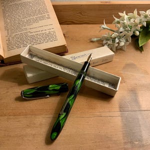 Może przedstawiać: Zabytkowe pi&oacute;ro wieczne z czarno-zielonym marmurkowym wzorem, wraz z oryginalnym pudełkiem. Pi&oacute;ro jest otwarte, odsłaniając stal&oacute;wkę, a skuwka jest umieszczona w pobliżu. Otwarta książka i białe kwiaty w tle.