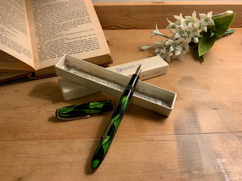Może przedstawiać: Zabytkowe pi&oacute;ro wieczne z czarno-zielonym marmurkowym wzorem, wraz z oryginalnym pudełkiem. Pi&oacute;ro jest otwarte, odsłaniając stal&oacute;wkę, a skuwka jest umieszczona w pobliżu. Otwarta książka i białe kwiaty w tle.