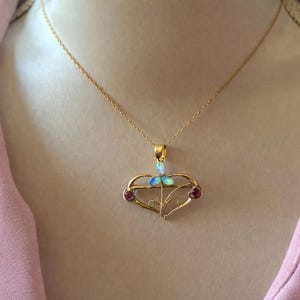 May include: A gold pendant necklace with a heart-shaped design. The pendant features an opal and two red gemstones. The necklace is worn against a pink background.
