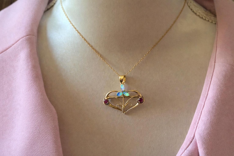 May include: A gold pendant necklace with a heart-shaped design. The pendant features an opal and two red gemstones. The necklace is worn against a pink background.
