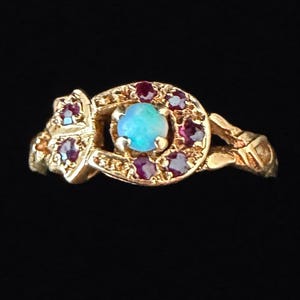Könnte beinhalten: Ein Goldring mit einem zentralen, runden Cabochon-Opal mit blauem und grünem Farbspiel. Der Opal ist von einer hufeisenförmigen Fassung umgeben, die mit kleinen, runden, tiefroten Rubinen besetzt ist. Die Ringschiene weist dekorative Metallarbeiten auf.