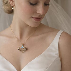 May include: A gold necklace with a heart-shaped pendant featuring a light blue butterfly and two dark red gems. The necklace is worn by a person wearing a white dress and veil, with a pearl earring.