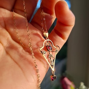 Puede incluir: Un collar con colgante dorado con un diseño geométrico y piedras preciosas rojas. El colgante tiene una forma de diamante central con una piedra preciosa más pequeña en forma de lágrima colgando debajo. El collar se sostiene en una mano.