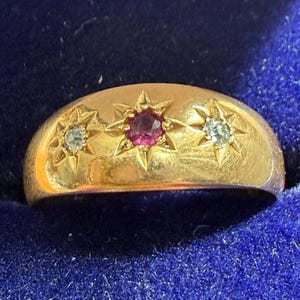 Puede incluir: Un anillo de oro con un diseño abovedado, con una gema roja central y dos gemas claras más pequeñas. Cada gema está engastada en un diseño de estrella. El anillo se muestra sobre una superficie azul.