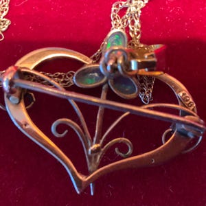 May include: A gold-coloured heart-shaped brooch with a floral design and a pin fastening. The brooch features a small flower with iridescent stones and is attached to a delicate chain. The number "1961" is stamped on the side of the heart.