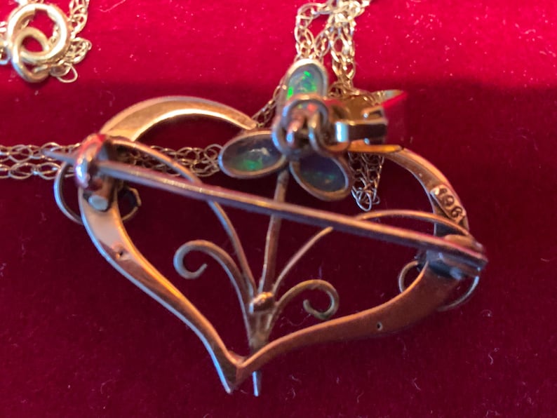 May include: A gold-coloured heart-shaped brooch with a floral design and a pin fastening. The brooch features a small flower with iridescent stones and is attached to a delicate chain. The number "1961" is stamped on the side of the heart.