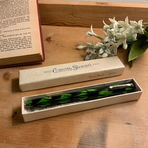 Może przedstawiać: Biały ceramiczny kubek z czarnym uchwytem i napisem "But First, Coffee" pogrubioną, czarną czcionką. Kubek jest ustawiony na rozmytym tle, sugerującym przytulne otoczenie.