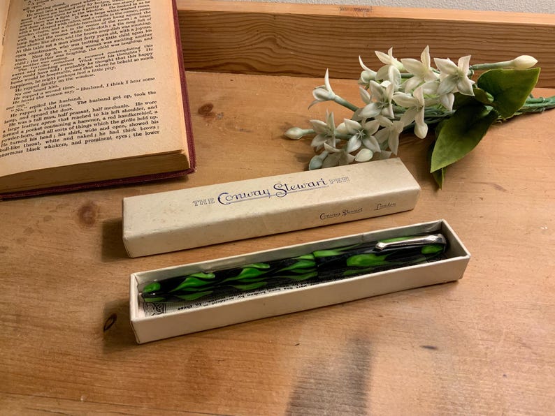 Może przedstawiać: Biały ceramiczny kubek z czarnym uchwytem i napisem "But First, Coffee" pogrubioną, czarną czcionką. Kubek jest ustawiony na rozmytym tle, sugerującym przytulne otoczenie.