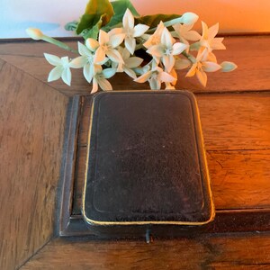 May include: A closed, rectangular black box with gold edging, placed on a wooden surface. White flowers with green stems are arranged above the box. The box is likely a jewellery or trinket case.
