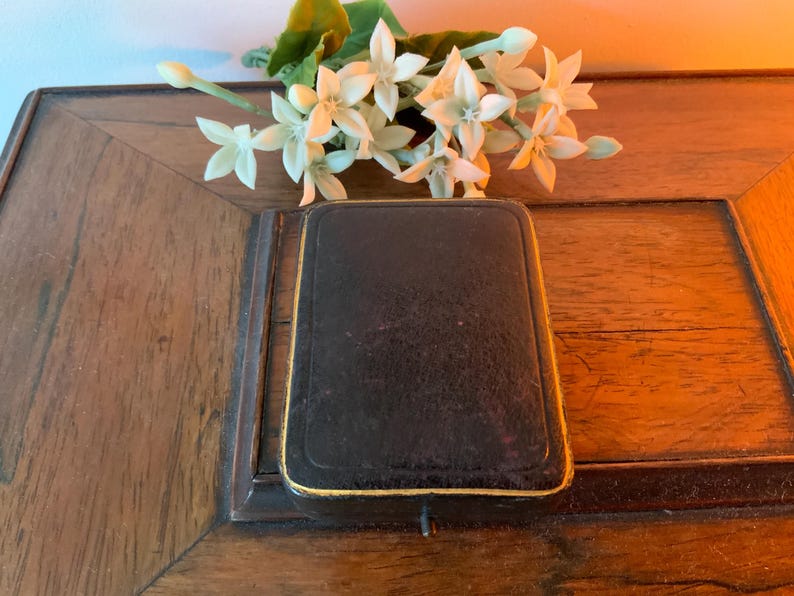 May include: A closed, rectangular black box with gold edging, placed on a wooden surface. White flowers with green stems are arranged above the box. The box is likely a jewellery or trinket case.