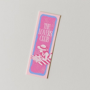 May include: A rectangular bookmark with a light pink background featuring a scalloped blue border. The center displays a pink graphic of a person wearing a sun hat and reading a book on a striped towel, with the text THE LOVERS CLUB in white.