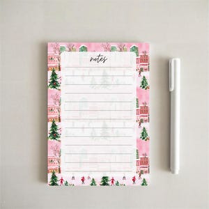 May include: A rectangular notepad with a pink winter scene border featuring watercolour illustrations of evergreen trees, buildings, and people skating, with the word notes printed at the top above horizontal lines, accompanied by a white pen.