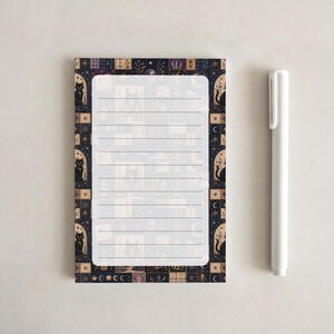 May include: A rectangular notepad with horizontal lines and a dark border featuring black cats, crescent moons, and celestial patterns, accompanied by a plain white plastic pen. The notepad has a light gray center with faint illustrations.