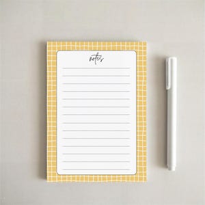 May include: A rectangular notepad with a yellow and white grid pattern border and a white center featuring horizontal lines and the word notes printed in black script at the top, accompanied by a plain white cylindrical pen positioned to the right.