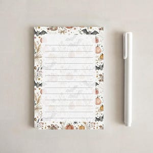 May include: A rectangular notepad with horizontal lines and a border featuring watercolor illustrations of bats, pumpkins, dried flowers, and spiderwebs, accompanied by a plain white plastic pen positioned vertically to the right side.