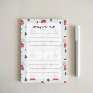 May include: A rectangular notepad with a light blue background featuring a pattern of gray cats in red sweaters, Santa hats, and green pine trees. The center has a white lined area with the text meowy christmas at the top, next to a white pen.