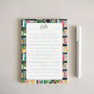 May include: A rectangular notepad with a white center featuring horizontal lines and the word Notes in black script. The border displays a colorful illustration of bookshelves filled with various books and potted plants. A white pen sits nearby.