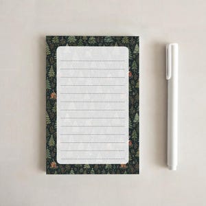 May include: A rectangular notepad with a dark green border featuring a pattern of small trees and houses, surrounding a white center with horizontal lines. A plain white pen is positioned vertically to the right of the notepad.