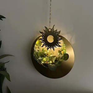 Może przedstawiać: Wisząca, okrągła, złota metalowa donica z wycięciem w kształcie słońca u góry. Donica jest wypełniona zielenią i małymi białymi światełkami. Łańcuch zawiesza donicę od góry.
