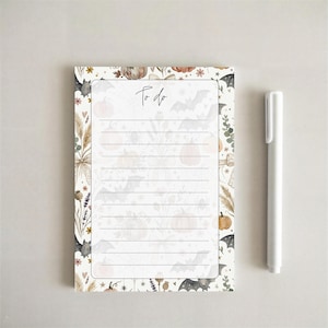 May include: A rectangular to-do notepad featuring a white background with a border of pumpkins, bats, spiders, and dried flowers. The center contains horizontal lines for writing and the text To do at the top. A white pen is positioned to the right.