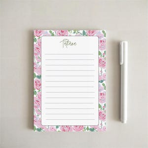 May include: A lined notepad with a floral rose design border and the name "Patiana" in cursive script. A white pen rests beside the notepad, ready for use. The notepad is approximately 8 inches tall and 5 inches wide.