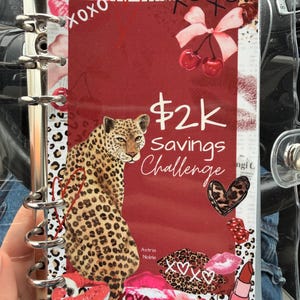 May include: A clear binder with a red cover featuring a leopard, lipstick, cherries, and the text "$2k Savings Challenge." The design includes kiss marks, hearts, and the words "XOXO."
