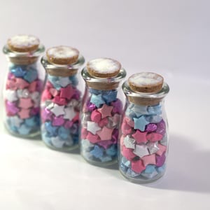 Peut inclure: Quatre bouteilles en verre transparent remplies d'étoiles en origami colorées. Les étoiles sont roses, bleues, blanches et argentées. Chaque bouteille est bouchée par un bouchon en liège.