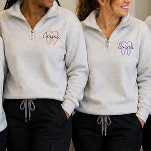 Puede incluir: Un grupo de personas con pul&oacute;veres grises claros con cremallera de cuarto de cierre, dise&ntilde;os de dientes bordados y nombres. Los pul&oacute;veres tienen un peque&ntilde;o bolsillo y se combinan con pantalones negros con cord&oacute;n. Los dise&ntilde;os de dientes son de varios colores.