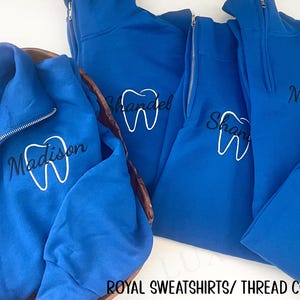 Puede incluir: Sudaderas azul real con cremallera de cuarto de cremallera y bordados blancos de dientes y nombres. Las sudaderas tienen una cremallera plateada y se muestran sobre una superficie blanca. El texto "ROYAL SWEATSHIRTS/ THREAD COLORS: BLACK & WHITE" est&aacute; en la parte inferior.