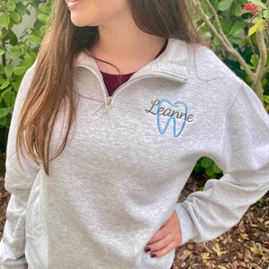 Puede incluir: Sudadera gris claro con cremallera de cuarto de cremallera, bordada con el nombre "Leanne" y un dise&ntilde;o de diente azul. La sudadera tiene una cremallera plateada y se usa sobre una camisa burdeos.