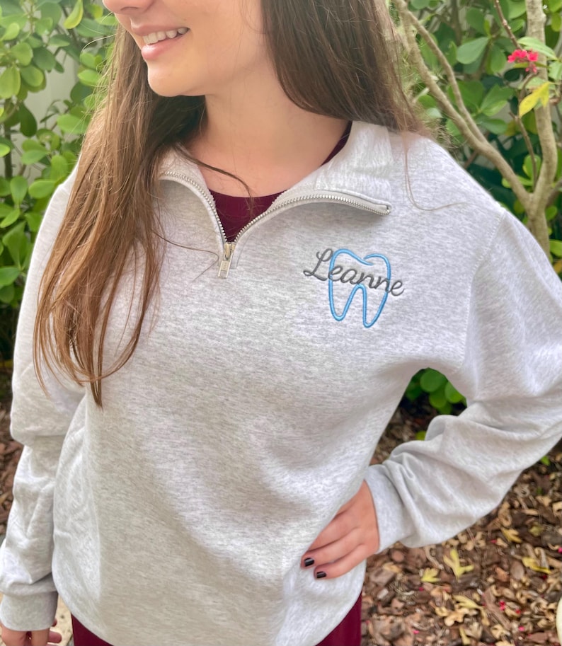Puede incluir: Sudadera gris claro con cremallera de cuarto de cremallera, bordada con el nombre "Leanne" y un dise&ntilde;o de diente azul. La sudadera tiene una cremallera plateada y se usa sobre una camisa burdeos.