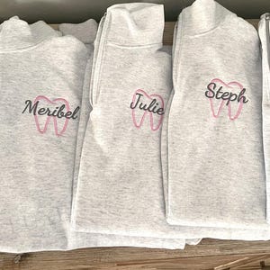Puede incluir: Se muestran cinco sudaderas grises claras con cuello con cremallera. Cada sudadera tiene un nombre bordado en escritura negra sobre un gr&aacute;fico de diente rosa. Los nombres son Kassandra, Meribel, Julie, Steph y Amanda.