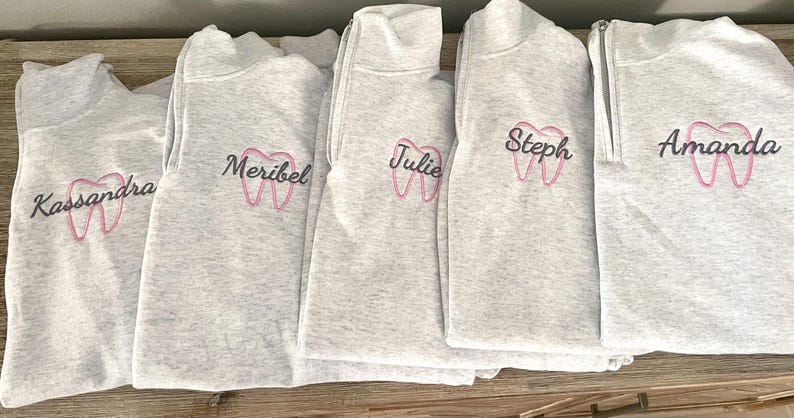 Puede incluir: Se muestran cinco sudaderas grises claras con cuello con cremallera. Cada sudadera tiene un nombre bordado en escritura negra sobre un gr&aacute;fico de diente rosa. Los nombres son Kassandra, Meribel, Julie, Steph y Amanda.