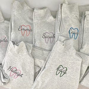 Puede incluir: Un conjunto de sudaderas grises con cremallera de cuarto de cierre, bordadas con dise&ntilde;os de dientes y nombres. Los nombres y los dientes est&aacute;n bordados en varios colores, incluyendo rosa, azul y verde. Las sudaderas tienen cuello alto.