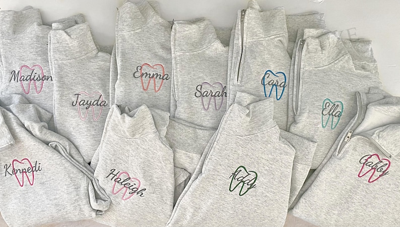 Puede incluir: Un conjunto de sudaderas grises con cremallera de cuarto de cierre, bordadas con dise&ntilde;os de dientes y nombres. Los nombres y los dientes est&aacute;n bordados en varios colores, incluyendo rosa, azul y verde. Las sudaderas tienen cuello alto.