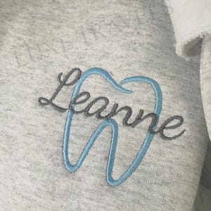 Puede incluir: Sudadera gris claro con un dise&ntilde;o bordado. El dise&ntilde;o presenta el nombre "Leanne" en escritura gris, con la letra "e" formando el contorno de un diente en azul claro. El texto "THREAD COLORS: CIEL BLUE & GRAY" est&aacute; en la parte inferior.