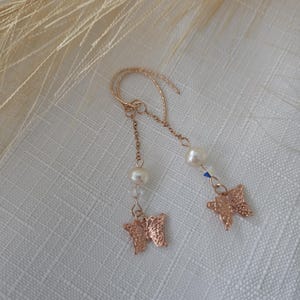Könnte beinhalten: Ein Paar roségoldfarbene Ohrhänger mit feinen Ketten, die jeweils mit einer weißen Perle und einem kleinen, detaillierten Schmetterlingsanhänger am unteren Ende verziert sind. Die Ohrringe sind mit geschwungenen Ohrhaken ausgestattet.