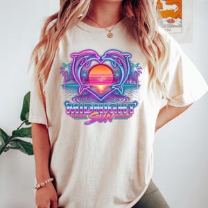 May include: Cream-colored t-shirt featuring a neon graphic of two dolphins forming a heart around a sunset. The words "MIDNIGHT SUN" are displayed below the graphic in a retro font. The shirt has short sleeves and a relaxed fit.