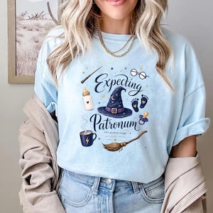 May include: A light blue t-shirt featuring a graphic print with the text "Expecting Patronum" and "Our greatest magic arrives soon." The design includes a wizard hat, a baby bottle, round glasses, baby footprints, a pacifier, and a broom.
