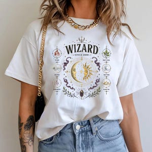 May include: A white short-sleeve t-shirt featuring a graphic print with the text WIZARD SINCE 2008. The design includes a central sun and moon illustration surrounded by smaller circular icons labeled Fire, Nature, Wind, Water, Energy, and Earth.