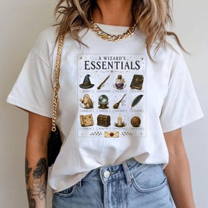 May include: A white t-shirt featuring a graphic print titled A WIZARD'S ESSENTIALS. The design displays a grid of twelve magical items including a hat, wand, potion, book, owl, orb, broom, feather, map, chest, candle, and ball with labels below each.