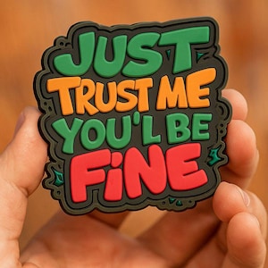 Puede incluir: Un parche rectangular colorido con las palabras "JUST TRUST ME YOU'LL BE FINE" en verde, naranja y rojo. El texto está delineado en negro y tiene un borde decorativo. El parche está en una mano.