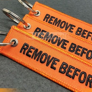 Puede incluir: Primer plano de tres etiquetas de tela naranja con el texto "REMOVE BEFORE" bordado en negro. Cada etiqueta tiene un llavero de metal plateado. Las etiquetas son probablemente para uso aeronáutico o industrial.